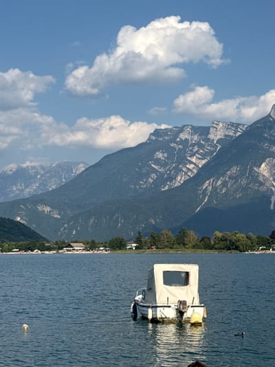 The image depicts a scenic view of Calceranica al Lago, Italy, on a clear day with a blue sky dotted with white, puffy clouds. In the foreground, a calm, blue lake occupies the majority of the frame, reflecting the light. A small, white motorboat with a beige canopy is moored in the middle of the lake, facing away from the camera. The boat has a visible outboard motor and a yellow buoy is attached to its stern. To the left of the boat, a small, round, yellow object floats on the water. In the distance, the shoreline is lined with trees and buildings, including what appears to be a resort or residential area. Behind the shoreline, a range of majestic, dark green mountains rises, their rugged peaks illuminated by the sun. The overall impression is one of a peaceful and picturesque summer day in a lakeside town.
