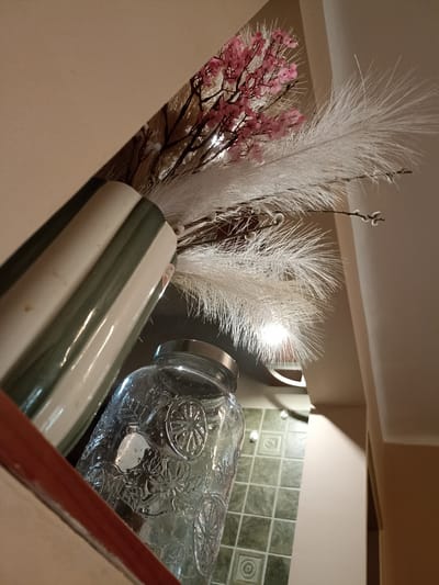 The image shows a still life composition within an indoor setting. In the foreground, a clear glass jar with decorative embossed patterns sits prominently. Behind the jar, a vase with green and off-white vertical stripes contains a floral arrangement. The arrangement includes delicate pink blossoms on dark branches and large, fluffy white plumes. The background reveals a tiled floor and a beige wall. The lighting suggests an indoor environment, possibly a home or a decorative display area. There is no indication of the location in Sosnowiec, Poland, or any people present in the image.