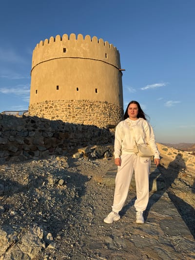 A female subject stands centrally positioned in front of the Hatta Fort watchtower in Hatta, United Arab Emirates. The individual, appearing as a young adult with long dark hair, wears a complete white athletic ensemble consisting of a hooded sweatshirt with subtle horizontal stripe detailing, sweatpants bearing a "Hollister" logo on the left thigh, and white athletic footwear. A light-colored crossbody bag, featuring a smaller attached pouch, is slung over her right shoulder. She maintains a frontal pose towards the camera, exhibiting a slight smile. The multi-tiered, circular watchtower, characterized by a rough stone base and a smooth, light-brown upper section with crenellated battlements and small rectangular apertures, occupies the background. A single wooden beam protrudes horizontally from the upper right quadrant of the tower. The scene is illuminated by low-angle sunlight, casting extended shadows from the left across the uneven, rocky, and gravel-covered terrain. The sky above is clear and blue with minimal cloud presence.
