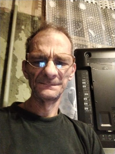 This is a selfie of a man in his late 50s or early 60s, with fair skin and thinning brown hair. He is wearing glasses with thin, gold-colored wire frames and a dark green t-shirt. The man is positioned in the center of the frame, with his head and shoulders visible. He has a weathered face with wrinkles and stubble. His expression is neutral.

Behind the man, a dark-colored television is visible, with its back facing the camera. The television has a grid pattern of ventilation holes and several ports labeled with icons and text, including what appear to be HDMI ports. To the left of the television, a white, patterned curtain is visible, and further to the left, a wall with peeling paint is present. The lighting in the image suggests an indoor setting, possibly a home, and the overall impression is informal. There is no clear indication of the time of day or weather. No text other than the labels on the television is legible. The setting is identified as Noginsk, Russia.