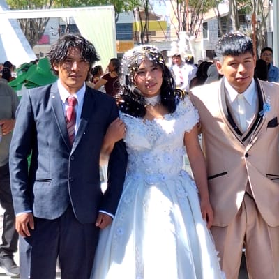 A medium shot captures a bride and groom, potentially after a wedding ceremony, in what appears to be Cerro Colorado, Peru. The bride wears a white wedding dress with floral accents. Her hair is adorned with decorative pieces. The groom wears a dark suit with a red tie. They stand outdoors, and additional people and structures are visible in the background, suggesting an urban environment.