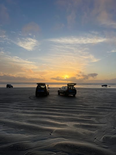Three SUVs are parked on a beach at sunset. The sky is a mix of blue and orange hues, with clouds scattered across the horizon. The sand is textured with tire tracks and natural patterns. The SUVs are facing in the same direction, and one has its trunk open. The overall scene evokes a sense of tranquility and adventure.