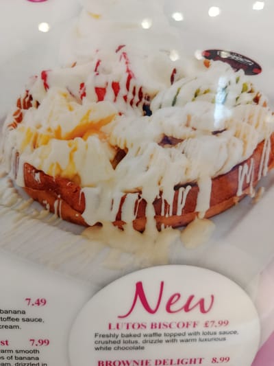The image shows a close-up of a dessert, likely a waffle, covered in white icing and drizzled with red sauce. The dessert is presented on a white plate, and the background is blurry, suggesting it is an indoor setting, possibly a cafe or restaurant. Text is visible in the lower right portion of the image, with the word "New" prominently displayed, followed by "LOTUS BISCOFF £7.99" and a description of the dessert. Other text below describes "BROWNIe DELIGHT 8.99". This context suggests the image is from a menu or promotional material in a food establishment. There are no people or specific landmarks visible to indicate a location in Bedford, United Kingdom. The focus is solely on the dessert and its description.