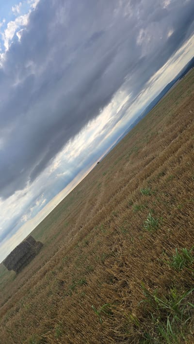 The image shows a field with harvested crops, likely wheat or barley, in Slatina, Bulgaria. The field is mostly covered in stubble with some patches of green grass. In the foreground, there are two rectangular bales of hay stacked on top of each other. The sky above is partly cloudy with dramatic dark clouds interspersed with shafts of sunlight. In the distance, rolling hills can be seen under the sky. A single utility pole is visible in the middle ground of the field. The perspective is angled, with the field stretching diagonally across the frame from the lower left to the upper right.