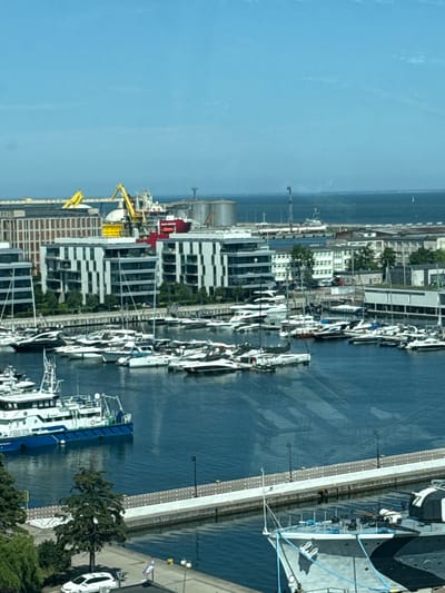 The image depicts a wide shot of a bustling harbor in Gdynia, Poland, likely taken from an elevated position. The scene is dominated by a deep blue body of water filled with numerous boats and yachts of varying sizes. In the foreground, a large, grey and black camouflaged ship with a prominent gun turret is docked. Further out in the harbor, a flotilla of smaller white yachts and boats are neatly arranged.

Beyond the marina, a modern waterfront development with multi-story buildings featuring white facades and numerous windows can be seen. These buildings appear to be residential or commercial. In the background, industrial elements of the port are visible, including large silos, cranes, and a large cargo ship. The sky is a clear, bright blue with a few wispy clouds.

The overall impression is one of a vibrant port city where maritime activity and modern urban living coexist. The sunlight casts reflections on the water, creating a dynamic visual texture. The scene is clean and orderly, suggesting a well-maintained harbor area.