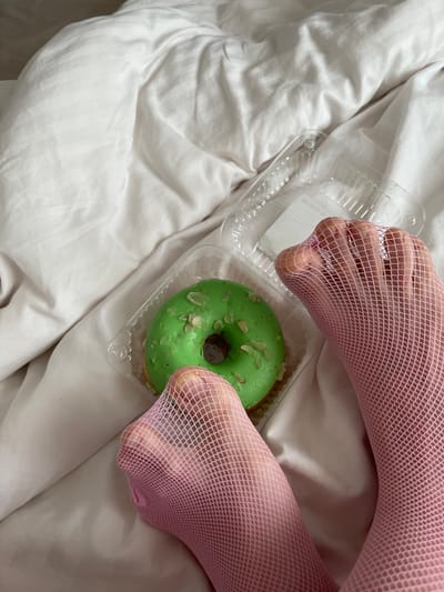 A close-up shot shows a green donut with white slivers of nuts inside a clear plastic container. The container is placed on a rumpled white sheet. Two feet, covered in pink fishnet stockings, are positioned on either side of the donut. The toes of the feet are visible through the mesh. The lighting is soft, suggesting an indoor setting with diffused light, possibly natural light from a window or soft artificial lighting. The overall impression is intimate and perhaps a bit playful, with the contrasting textures of the stocking, the sheet, and the donut creating visual interest. The focus is on the feet and the donut, implying a personal moment or a specific aesthetic. The scene is likely set in a bedroom or a similar private space, given the presence of the sheet. There is no visible text in the image.
