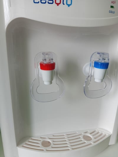 This media file shows a close-up, eye-level shot of a white water cooler. The water cooler has two dispensers, one for hot water and one for cold water. The hot water dispenser has a red cap, and the cold water dispenser has a blue cap. Both dispensers have clear plastic shields over them. Above the dispensers, the word "HOT" is printed in red text with a green indicator light next to it, and the word "COLD" is printed in blue text. The water cooler is sitting on a white drip tray with horizontal slots. The background is a plain, light-colored surface. The scene appears to be indoors, possibly in a kitchen or office. There are no people visible in the media file. The lighting is bright and even, suggesting it was taken during the day. The visible text is "HOT" and "COLD" along with the brand name "CESUQL" at the top of the water cooler.