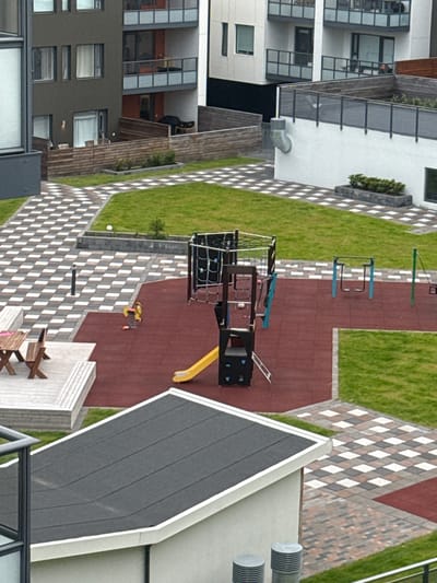 This is a high-angle, full-shot photograph of a modern apartment complex courtyard in Reykjavik, Iceland. The courtyard features a playground area with red rubber surfacing, a climbing structure with nets, a yellow slide, and a red rocking horse. Adjacent to the playground is a paved area with a checkered pattern of light and dark gray and white tiles, bordered by green grass. A wooden picnic table and chairs are situated on a raised wooden deck. In the background, buildings with balconies and dark windows are visible, with wooden fences and planters scattered throughout the outdoor space. The weather appears to be overcast.