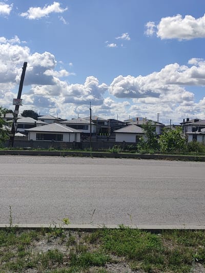 The image captures a residential area under a partly cloudy sky. In the foreground, a grey asphalt road runs horizontally, bordered by a concrete curb and overgrown grass and weeds. Behind the curb, a row of houses with modern architecture and grey tiled roofs are visible. These houses appear to be newly constructed and are separated by low, grey brick walls and fences. Some of the houses have dark-rimmed windows. A wooden utility pole stands to the left of the frame, leaning slightly, with a small sign attached to it featuring Cyrillic text. Green trees and bushes are interspersed between the houses and along the property lines. The overall impression is that of a suburban or newly developed neighborhood. The scene is located in Velykyi Zhytyn, Ukraine.