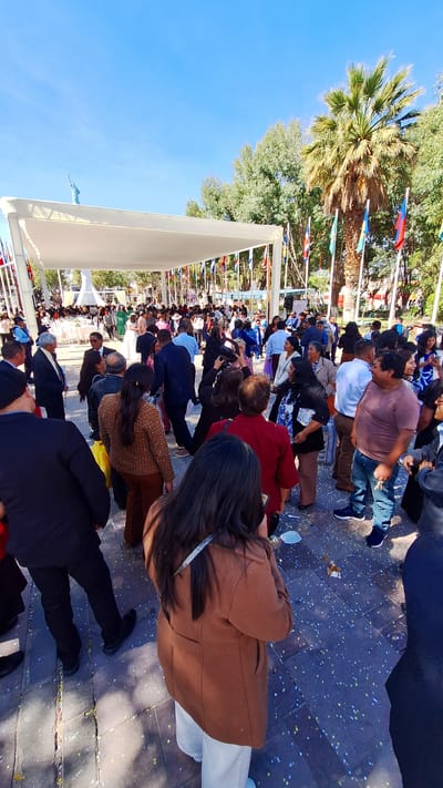 In Cerro Colorado, Peru, a large crowd is assembled outdoors on a paved area, possibly a town square, on a sunny day. People of various ages and attire are present, suggesting a public event. Confetti litters the ground. A canopy is erected over a portion of the crowd. Flags from numerous countries are displayed along the perimeter of the event. Trees, including a palm tree, are visible.