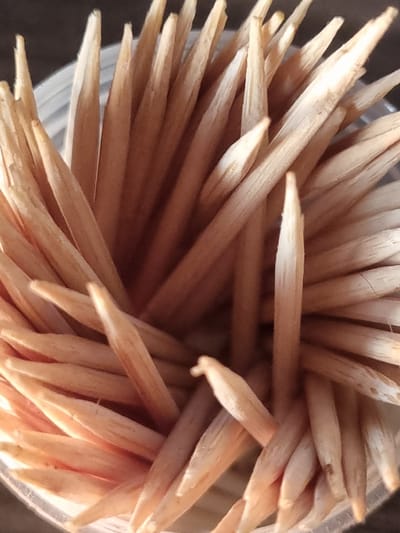 The image is a close-up, overhead shot of a bundle of wooden toothpicks. The toothpicks are tightly packed together, with their sharpened ends pointing upwards. The lighting is natural and appears to be coming from above, creating highlights and shadows on the textured surfaces of the wood. The overall color palette is warm, dominated by natural wood tones ranging from light beige to a deeper brown. The background is dark and out of focus, drawing all attention to the toothpicks. There is no evidence of any specific location in Žygaičiai, Lithuania, or any other geographical context. The image focuses solely on the arrangement and texture of the toothpicks.