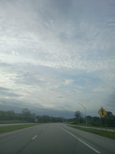 The image shows a highway scene with a cloudy sky and trees in the background. A yellow diamond-shaped sign with a black arrow pointing upwards and a line branching off to the left is visible on the right side of the road. There are streetlights along the highway. The road has clear lane markings, indicating it is a multi-lane highway. The general appearance suggests a rural or semi-rural setting outside of a major city. The location is identified as Winchester, United States.