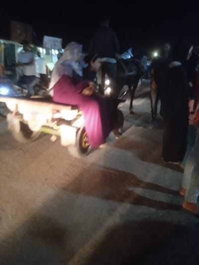 This image depicts a nighttime scene in Al-Mawasi, Palestinian Territories. In the foreground, a person is seated or lying on a motorized, two-wheeled cart, draped in a bright purple garment and appearing slumped forward. A light source emanates from the front of the cart, illuminating the immediate area. Behind the cart, another individual in a light-colored garment is partially visible. In the mid-ground to the right, a dark-colored horse or donkey is being ridden by a person. Multiple indistinct human figures are present in the background and the right side of the foreground, suggesting a gathering or movement of people. The ground is a dark, unpaved or poorly lit surface, and long shadows are cast by individuals and objects, indicating multiple light sources within the scene.