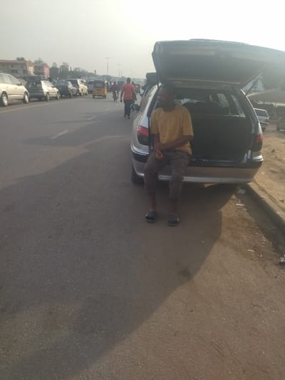 A man wearing a yellow shirt and brown pants sits in the open trunk of a silver car.  The car is parked on the side of a paved road with traffic in the distance.  The man is looking towards the right side of the image.  The location, at latitude 9.024, longitude 7.573, is a street in Maraba, Nigeria.  The man's feet are bare and he is wearing flip flops.  The car's trunk is open and the interior is visible.  There are multiple vehicles parked on the roadside and traffic is moving in the distance.  The image is taken in a sunny daytime setting with a hazy sky.