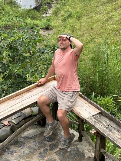 A person is standing on a weathered wooden structure in Yên Minh Commune, Vietnam, likely a viewing platform or elevated walkway. The person has a beard and dark hair, wearing a pink short-sleeved shirt, tan shorts, and gray sneakers. Their right hand is raised to their forehead, shielding their eyes. The background includes dense green vegetation, including trees and tall grasses, suggesting a natural environment. The wooden structure is built over a textured stone or tile surface.
