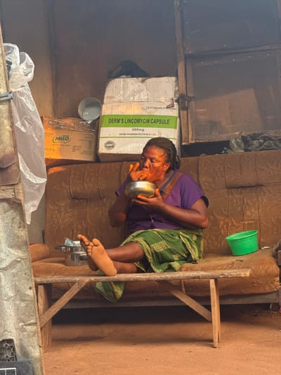 An older woman in a purple shirt and green patterned skirt sits on a wooden stool, holding a silver bowl to her lips. The scene appears to be indoors, with various boxes stacked behind her. One box is labeled "DERM'S LINCOMYCIN CAPSULE 500mg," and another is labeled "MILO." A green bucket hangs on the wall nearby. The background shows simple construction.