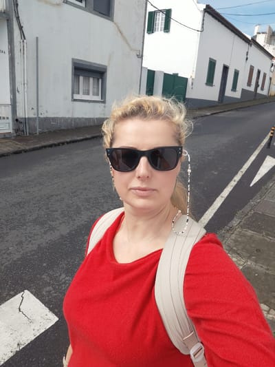 A woman with blonde hair and wearing sunglasses stands on a street in Ponta Delgada, Portugal. She is wearing a red shirt and a tan backpack. The street is paved and slightly inclined. There are white buildings with gray trim and dark green or brown shutters on either side of the street. There are crosswalk markings visible on the street.