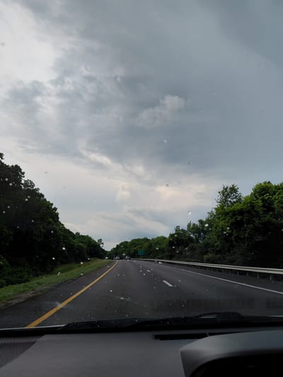The image shows a view from inside a vehicle traveling on a highway, likely near Frankfort, United States of America. The sky is heavily clouded, with a grey, overcast appearance. There are visible water droplets on the windshield. On either side of the highway are dense green trees. The highway itself is paved, with a yellow line marking the left lane and white lines marking the lanes. A guardrail is visible on the right side of the road. In the distance, there are a few other vehicles on the highway and a green sign indicating directions to various locations. The overall scene suggests a cloudy or possibly rainy day.