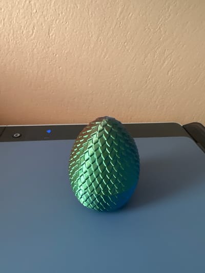 A metallic, green, dragon-egg-shaped object is centered on a flat, blue surface. The object has a textured, scale-like pattern. Behind the object, the top edge of a black electronic device is visible, with a small blue light emitting from it. The background consists of a textured, beige wall. The scene is an indoor setting, and the specific location in Hradec Králové, Czechia, cannot be determined from the image.
