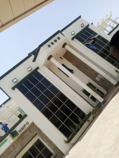 The image shows a modern building with a white facade and dark-tinted windows. The building has a prominent entrance supported by white columns and decorative stone accents. The NNPC logo is visible on the side of the building. A person in a blue uniform is seen near the entrance, possibly performing maintenance or cleaning. Several potted plants are placed in front of the building, adding greenery to the scene. The sky is clear and bright, suggesting it is daytime. The overall setting appears to be an office building or a corporate facility in Hotoro North, Nigeria.