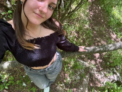 A young woman is sitting on a fallen tree trunk in an outdoor, wooded area, possibly in Lipnitsa, Bulgaria. She is wearing gray jeans, a black top with a ruffled texture on the upper portion, and a long-sleeved black jacket or blouse. She is also wearing white sneakers. The woman has long brown hair, fair skin, and is facing the camera. The area around the tree trunk is covered in grass, small flowering plants, and leaf litter. The sun is shining through the trees, casting shadows on the ground and the tree trunk. The scene appears to be natural, with rocks, vegetation, and soil in the background.