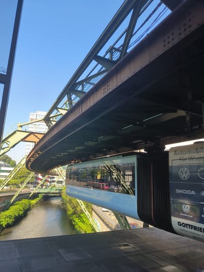 This is a medium-angle shot looking up at the Wuppertal Schwebebahn, a suspension railway in Wuppertal, Germany. The monorail train, painted a light blue, is suspended from a steel track and runs above a river lined with lush green trees. The sky is a clear, bright blue, suggesting a sunny day.

In the background, several buildings are visible, including a multi-story structure with many windows. A road runs alongside the river, with some cars and what appears to be a red bus visible. The infrastructure supporting the Schwebebahn is a prominent feature, with its large metal beams and supports dominating the upper part of the frame. The train itself has large windows that reflect the surrounding environment.

On the side of the train or a structure adjacent to it, there is visible text, including "Automobil" and "für Wuppertal" (Automobile for Wuppertal). Below this, logos for Volkswagen (VW) and Skoda are present, along with the name "GOTTFRIED" and the text "Uhrenhändler" (Watch dealer). The scene appears to be bustling with activity, as evidenced by the traffic and the train in motion. The overall impression is one of an urban landscape integrated with an innovative public transportation system.