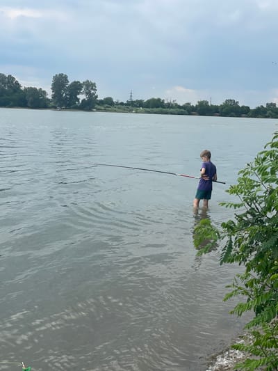 A young boy stands in the murky water of a river, facing away from the camera, holding a long fishing rod. He is wearing a dark purple t-shirt and teal shorts. The river is calm with small ripples on the surface. In the background, a line of trees separates the river from a cloudy sky. The location is Tliustenkhabl', Russia.