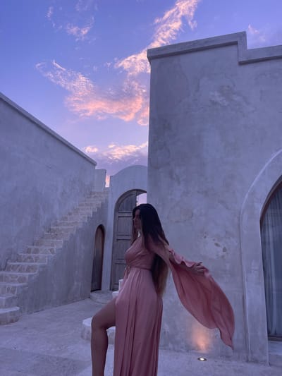 A woman in a pink dress stands in front of a building in an outdoor setting. The building is made of concrete and has stairs leading up to a doorway. The sky is purple and pink, with clouds. The woman is facing away from the camera, with her hair flowing behind her. She is posing with her arms outstretched. The setting appears to be a courtyard or an outdoor space. The image captures a moment of grace and movement against a backdrop of architectural elements and a colorful sky.