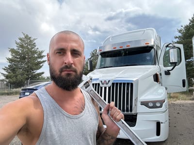 A man with a shaved head and beard, wearing a gray tank top, is taking a selfie in front of a white semi-truck. He is holding a large adjustable wrench in his right hand. The truck has its driver's side door open. In the background, there is a dark blue car, a chain-link fence, and pine trees under a cloudy sky. The scene appears to be a roadside or parking area.
