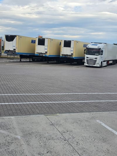The image shows a parked DAF truck with two yellow shipping containers stacked on its trailer. The truck is white, and the containers are light yellow with blue lettering that reads "NA". One container has a white refrigeration unit on its front. The truck and containers are parked in a lot paved with gray interlocking bricks, marked with white lines. To the right of the paved lot is a concrete surface. The sky is visible in the background, filled with white and gray clouds against a blue sky, suggesting it is daytime. The scene appears to be a parking area, possibly at a logistics or distribution hub, given the presence of the commercial truck and shipping containers. There is no visible activity or people in the image. The license plate of the truck is visible, reading "JDR 2464". Text on the containers and a sign on the truck's side are difficult to read clearly. The overall impression is one of a static scene in an industrial or commercial outdoor setting. The image is taken from a slightly elevated, angled perspective, rotated 90 degrees clockwise.