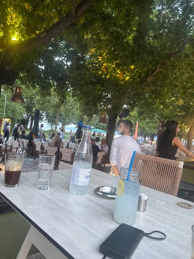 The image depicts an outdoor seating area of a cafe or restaurant in Asprovalta, Greece, likely during the late afternoon or early evening, given the lighting. In the foreground, a white marble-patterned table holds several drinks: a glass of dark iced coffee with foam, a glass of water, and a bottle of water. A glass jar with a pale blue beverage, a slice of lemon, and a blue straw is also on the table, alongside an ashtray and a small metal container. Further back, patrons are seated at tables under the shade of mature trees. A couple, a man in a white long-sleeved shirt and a woman in a black dress, are visible in the mid-ground, interacting with each other. Other people are seen socializing and dining at various tables. In the background, beyond the trees, a body of water and beachgoers suggest the cafe's proximity to the sea. The overall atmosphere appears relaxed and convivial, typical of a seaside resort town.