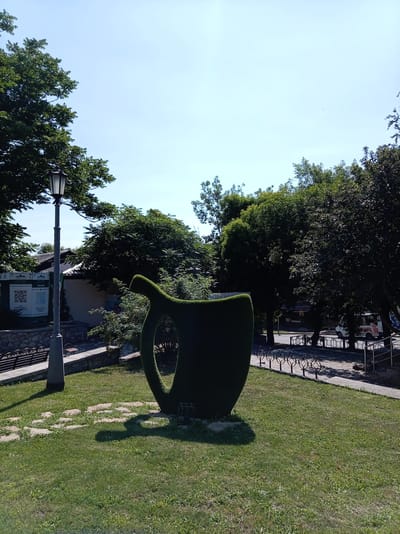 This is a daytime, eye-level, full exterior shot of a park area in the city of Zheleznovodsk, Russia. In the foreground, a large, dark green, topiary sculpture shaped like a teacup with a handle dominates the scene. The sculpture is casting a distinct shadow on the well-maintained green grass surrounding it. To the left of the sculpture, a tall, dark lamppost stands, with a bench and stone wall visible behind it. A path of stepping stones leads towards the sculpture. In the mid-ground, mature green trees form a dense backdrop, with some buildings and a street visible through the foliage. On the street, a white van is parked, and there are bicycle racks. The sky above is a clear, bright blue.