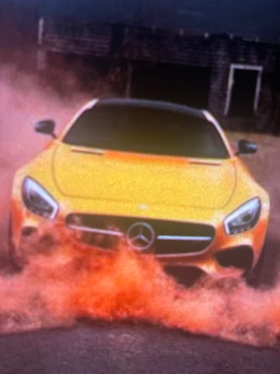 A yellow Mercedes-Benz sports car is pictured from the front, partially obscured by a cloud of vibrant orange smoke. The car is on a dark asphalt surface, and behind it, a dimly lit, rustic brick building with an open doorway is visible. The overall atmosphere of the image is dramatic and energetic, suggesting a moment of intense action or a staged performance. The location is identified as Paczkowo, Poland.