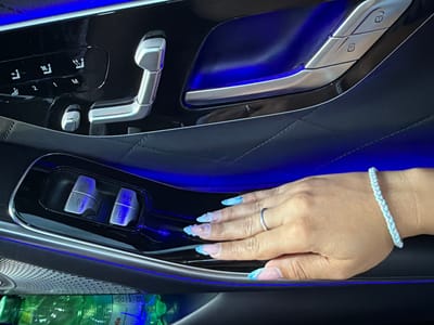 The image shows the interior of a car, likely a luxury vehicle, with blue ambient lighting. In the foreground, a person's hand with manicured nails is resting on the car's door panel. The hand is adorned with a braided bracelet and a ring. The door panel features various controls, including seat adjustment buttons and window controls. In the background, a partially visible bottle of water is seen. The image was taken in Kazachevo, Bulgaria.