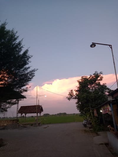 The image depicts a dusky, rural scene near Bungur, Indonesia. A paved road stretches into the distance, bordered by a rice field and various structures. On the left, a small open-sided pavilion with a tiled roof stands near a pile of gravel. A tall pine tree partially obscures this structure. On the right, a small store with a partial roof and hanging advertisements is visible, with a parked motorcycle nearby. A tall street lamp stands adjacent to the store. The sky is a mix of blue and pink hues with light pink clouds, suggesting either sunrise or sunset.