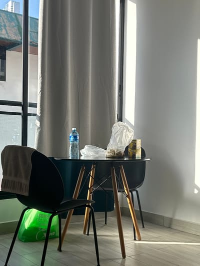 The image depicts an interior scene, likely an apartment or hotel room, in Panama City, Panama. A dark-colored, modern chair with a light-colored towel draped over its back is positioned in the foreground. Beneath the chair, a bright green reusable shopping bag with Spanish text is visible. A round table with a dark surface and wooden legs stands in the midground. On the table, there is a clear plastic bottle of water, a plastic bag containing some items, and two small boxes of Nesquik cereal. The room is lit by natural light streaming in from a window, casting distinct shadows on the white wall and tiled floor. A light-colored curtain hangs by the window. Outside the window, parts of a building and a green roof are visible.