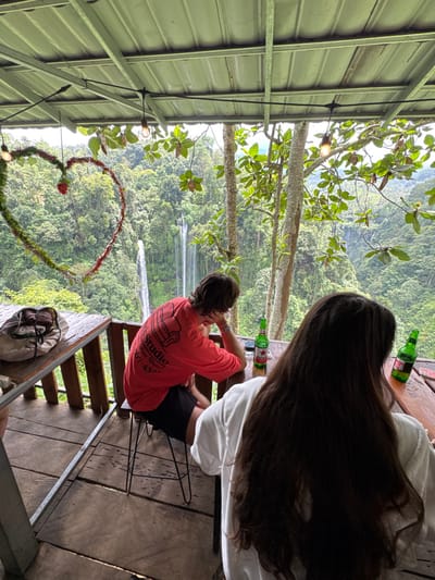 Two people sit at a wooden table on a deck, overlooking a lush green forest with a waterfall in the distance. A large heart-shaped decoration hangs to the left, and string lights are visible near the metal roof structure. Two green beer bottles sit on the table. One person wears a white shirt, and the other wears a red shirt with a logo. A bag rests on the wooden steps leading up to the deck.