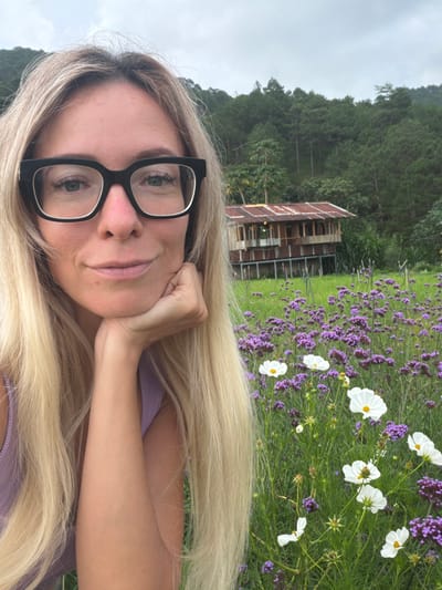 A blonde woman wearing black-rimmed glasses rests her chin on her hand, looking at the camera with a slight smile. Her hair is long and blonde, framing her face. She wears a light purple tank top. In the background, a field of purple and white flowers is visible, with a rustic wooden house and a pine forest on a hillside under a cloudy sky. The scene appears to be in Xuan Truong, Da Lat, Vietnam.