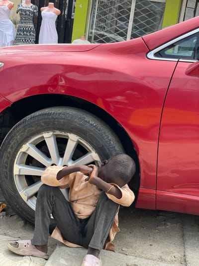 A young boy is seen sitting on the ground next to a red car. He is wearing a light brown shirt and dark grey pants, with a yellow band on his left wrist. His head is bowed and he appears to be resting, with his arms wrapped around his knees. The boy is wearing light pink sandals. The car has a visible tire and wheel. In the background, to the left of the car, there are mannequins dressed in white and patterned clothing, suggesting a shop or market. The ground is rough concrete. The scene appears to be outdoors, with what looks like natural light, possibly indicating daytime. The location is identified as Lagos, Nigeria.