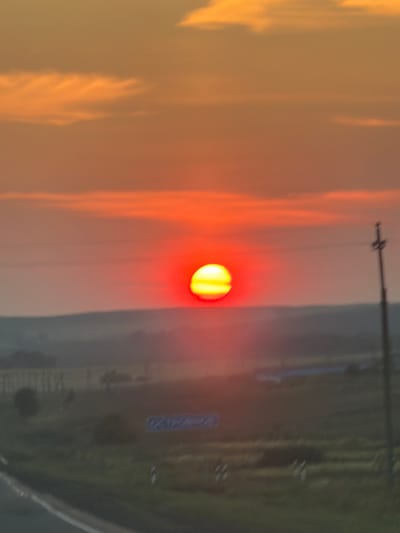 The image shows a blurry, low-angle view of a sunset over a rural landscape, likely in the city of Ostrovnoe, Russia. The sun is a bright, glowing orange orb, low on the horizon, surrounded by a hazy red and orange sky. Thin, wispy clouds are visible in the upper portion of the image. The middle ground consists of a dimly lit, undulating terrain with hints of vegetation and possibly some man-made structures. A dark, indistinct silhouette of trees or buildings is visible on the left. In the foreground, a blurred asphalt road with a white line is present. A single, dark utility pole stands on the right side of the frame, its crossbar indistinct. A faint blue sign with unreadable text is visible on the right side of the road. The overall impression is that of a scenic sunset in a relatively undeveloped area.