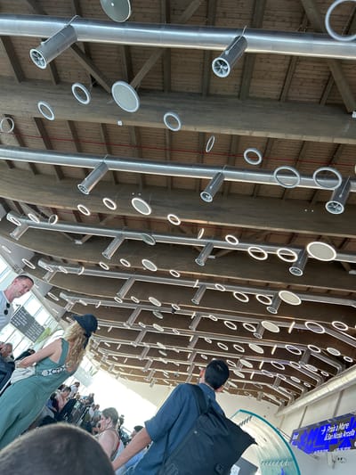 This media file captures an interior view of a modern, architecturally striking space, likely an airport terminal or a public transport hub in Lamezia Terme, Italy. The dominant features are the curved, wooden ceiling structure and a series of parallel, silver cylindrical ducts extending downwards. Suspended from these ducts are numerous circular elements, some appearing to be light fixtures with a white glow, and others possibly ventilation outlets with fan-like interiors. The overall aesthetic is industrial yet modern, with a focus on clean lines and metallic and wooden textures.

Several people are visible in the scene, suggesting it's a functional public space. In the foreground, a person wearing a blue shirt and a backpack is looking upwards, as if admiring the architecture. Further back, a woman with curly blonde hair and a green jumpsuit, wearing a dark cap, is also looking around, possibly waiting. Other individuals are seen in the background, some seated, others standing, hinting at a travel or transit scenario. The lighting suggests it's daytime, with natural light filtering in from the right side of the frame, creating bright highlights.

A notable detail is a blue digital display board in the upper right corner, partially visible. It shows text that appears to be route information or flight details. The legible text includes "→Praia a Mare & San Nicola Arcella" and "dal 26.06.2006 31.28.06.2006", indicating destinations and possibly dates related to travel. The presence of a digital display board and people with luggage (implied by the context and the backpack) strongly suggests this is an airport or train station. The clean, modern design and the open space evoke a sense of efficiency and contemporary travel.