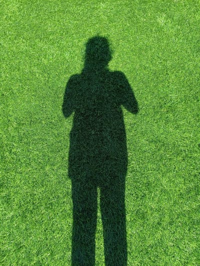 The image shows the shadow of a person casting on a bright green, short-cut lawn. The shadow is dark and clearly defined, indicating direct sunlight. The person's silhouette appears to be standing upright, with arms bent and held towards the chest. The texture of the grass is visible in detail, with individual blades discernible. The overall scene is outdoors, under bright lighting conditions. There is no indication of a specific location within Dnestrovsk, Moldova, or any urban elements in the image.