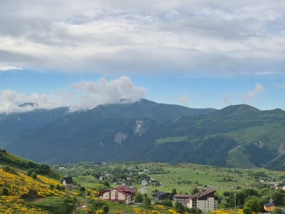 This is a scenic landscape photograph of Gudauri, Georgia, taken on a clear day with a mixture of sun and clouds. In the foreground, a grassy hillside is dotted with vibrant yellow flowers. Several buildings are visible, including a large hotel with a red roof and a smaller dark-colored building. A ski lift tower stands prominently in the mid-ground. Beyond the immediate hillside, a valley unfolds with more scattered houses and buildings, indicative of a village or resort town. The middle ground features rolling green hills and fields. The background is dominated by a majestic mountain range, its slopes covered in lush green vegetation. The sky is a bright blue, with scattered white and gray clouds creating a dynamic atmosphere. The overall impression is one of a peaceful, picturesque mountain setting in Gudauri.