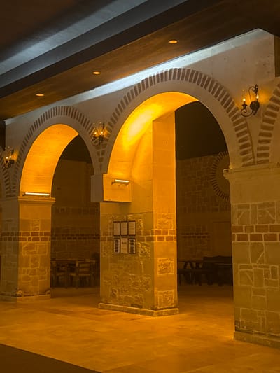 The image captures an architectural detail in what appears to be a building in Ürgüp, Turkey. It showcases a series of illuminated archways, constructed with light-colored stone and brick. The archways are lined with decorative bricks. There are dark voids that can be seen through the arches. In between the arches are stone pillars, with some featuring wall-mounted light fixtures. A set of small plaques are mounted on one of the pillars. The lighting casts a warm, golden hue across the structure. To the side of the arches are dark wooden beams.