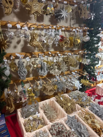 Rows of Christmas ornaments in gold, silver, and clear designs are displayed on wooden stands and in cushioned trays. The ornaments vary in shape and material, including stars, snowflakes, and delicate wire structures. Many are embellished with glitter or intricate cutouts. Greenery is placed on top and bottom of displays. Price tags dangle from the ornaments.