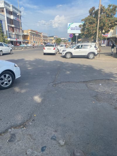The image shows a street scene in Parkent, Uzbekistan. In the foreground, the asphalt road has patches of damage and debris. To the left, the front quarter panel and wheel of a white car are visible. In the middle ground, several cars are driving or parked on the street, including a white sedan with the license plate "03 R 458 VA" and a white SUV. A large billboard with Uzbek text and green and white graphics is visible in the background, along with several multi-story buildings and trees. People can be seen walking and standing on the sidewalk and near the buildings. The sky is blue with scattered clouds. The overall impression is of a typical urban street on a sunny day.