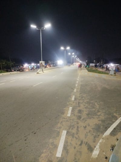 The image depicts a street scene at night in Akabuka, Nigeria. The street is illuminated by modern streetlights, with multiple lampposts lining the road, casting a bright glow. The road surface appears to be asphalt with white lane markings. On the right side of the road, there are several small structures that appear to be shops or stalls, with people gathered around them. Some of these structures are lit from within. On the left side of the road, there are also some buildings and trees visible. A few people are walking along the side of the road and in the distance. The overall impression is that of a moderately busy urban or semi-urban area at night.