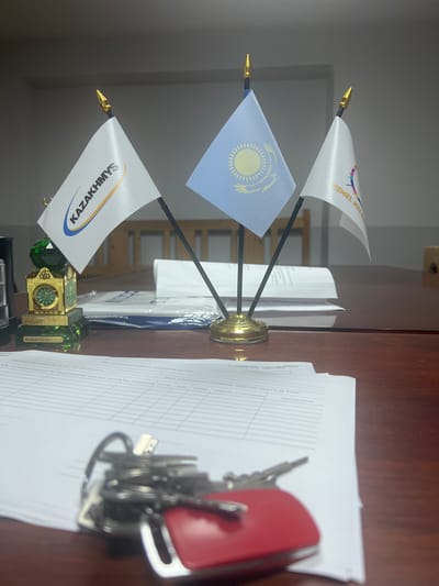 The image displays a desk with several objects, including three small flags on a gold stand. The flags feature the Kazakh flag, a white flag with "KAZAKHMYS" written in blue and gold, and a white flag with a colorful graphic and the words "KEMEL RELE." A small, ornate gold clock with a green crystal base is also visible. To the right of the flags are a stack of papers and a set of keys attached to a red key fob. The desk appears to be made of wood. The lighting suggests it is daytime, likely indoors in an office setting. No people are visible in the image. The text "KAZAKHMYS" and "KEMEL RELE" are clearly legible, along with some Cyrillic text on the papers. The presence of the Kazakh flag and the company name "KAZAKHMYS" suggests a connection to Kazakhstan.