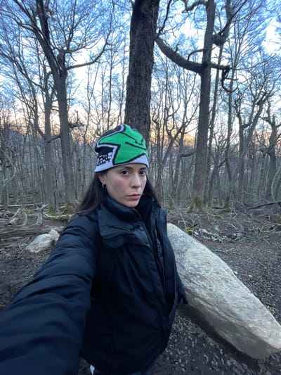 The image shows a woman with dark hair, wearing a green, black, and white beanie and a dark jacket, leaning against a large, pale, rectangular object that appears to be a rock or a large piece of wood. The background consists of a forest with many bare trees and branches, indicating it might be winter or a similar time of year when leaves have fallen. The trees are gray, and the sky is a pale blue or light. The ground is covered in leaves and debris.

Given the location of Unknown, Argentina, it is possible this area is a natural park or a forested area near the city.