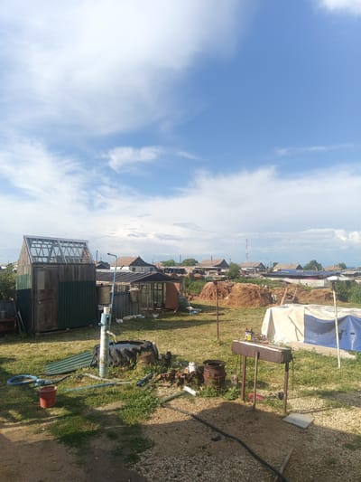 The image depicts a rural backyard scene in Ясная Поляна ауылдық округі, Kazakhstan. The setting is outdoors on a sunny day with scattered clouds in a blue sky. The ground is covered with grass and gravel.

Prominent objects in the scene include a greenhouse with a wooden frame and glass roof, a green-sided shed, a metal barbecue grill with a can of oil on top, and a rusty barrel. There is also a manual water pump with a blue handle, a large black tire, a red bucket, and various hoses and pipes scattered on the grass. In the background, several houses with pitched roofs are visible, along with stacks of hay or straw. A tall communication tower can be seen in the distance. A partially constructed or covered structure with blue and white tarps is in the middle ground. Some white chickens are also present on the grass near the shed.

The scene appears to be a residential property in a village, likely in the summer due to the greenery and sunny weather. No people are visible in the image. The overall impression is one of a functional, perhaps somewhat cluttered, outdoor living and working space in a rural environment. The light suggests it is daytime, likely mid-morning or afternoon. There is no visible text in the image.